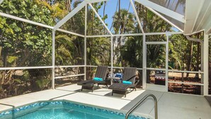 An indoor pool area with two lounge chairs and a small table, surrounded by glass walls and tropical plants.