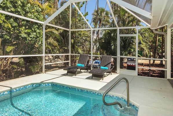 An indoor pool area with two lounge chairs and a small table, surrounded by glass walls and tropical plants.