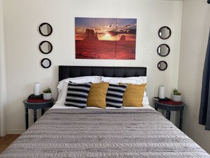 Una cama bien hecha con un edredón estampado, dos almohadas y un cuadro enmarcado encima. Hay dos mesitas de noche con espejos decorativos y pequeñas plantas.
