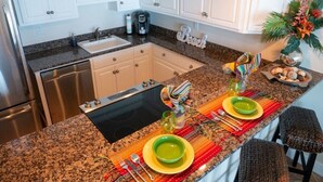 Une cuisine avec des meubles blancs, un plan de travail en granit, un évier, une machine à café et un panneau décoratif.