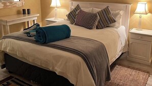 A neatly made bed with a rolled towel, two bedside lamps, a framed picture on the wall, and a small table with a mirror and two bottles.