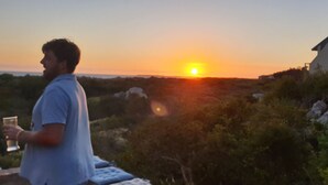 A person standing outdoors during sunset, holding a glass. There is a house in the background with a view of the sunset.