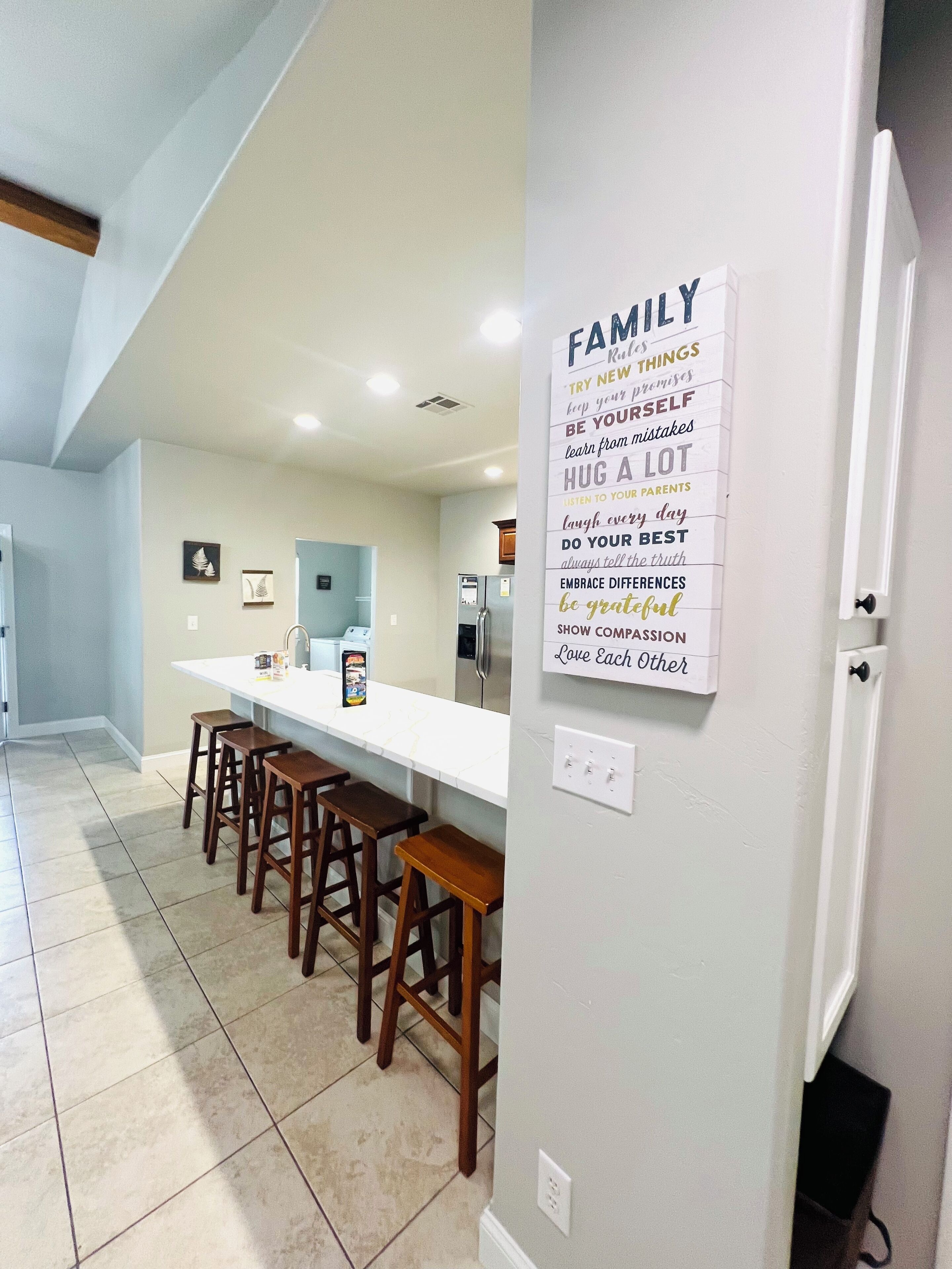 A modern kitchen with a long white countertop, wooden stools, and a wall-mounted sign with family values.