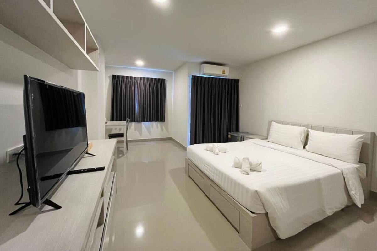 Una habitación de hotel moderna con televisor de pantalla plana, una cama con sábanas blancas, una mesita de noche y un equipo de aire acondicionado instalado en el techo.