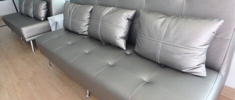 Un sofá moderno tapizado en gris con varios cojines, ubicado en una habitación amplia y luminosa con piso de baldosas.
