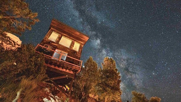 Una cabaña de madera con balcón, rodeada de árboles, bajo un cielo estrellado.