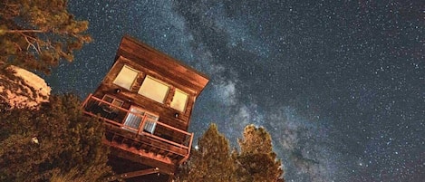 A wooden cabin with a balcony, surrounded by trees, under a starry night sky.