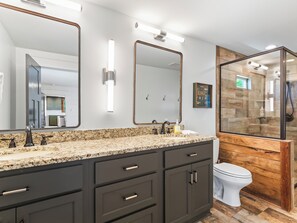 Un baño moderno con encimera de granito, muebles oscuros, inodoro blanco y zona de ducha con mampara de vidrio.