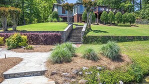 A modern house with a stone pathway leading to a wooden deck, surrounded by well-maintained lawns and various shrubs.
