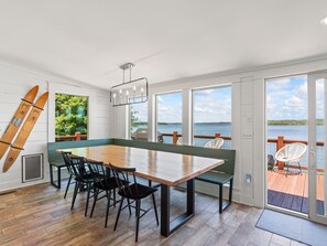 Un comedor con mesa y sillas de madera, ventanas amplias con vista al lago, y dos tablas de surf apoyadas contra la pared.
