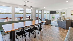 A spacious room with a large wooden table and chairs, a sofa, and a TV. It features large windows with a view of a lake and a deck area.