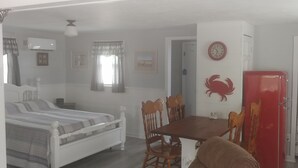 A bedroom with a bed, a dining table with chairs, a red refrigerator, and a wall clock. - Island Time Cottage (Chincoteague)