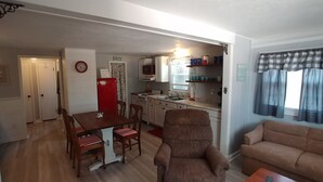 A living room with a brown sofa, a wooden dining table with chairs, a kitchen area with cabinets and appliances, and a red refrigerator. - Island Time Cottage (Chincoteague)
