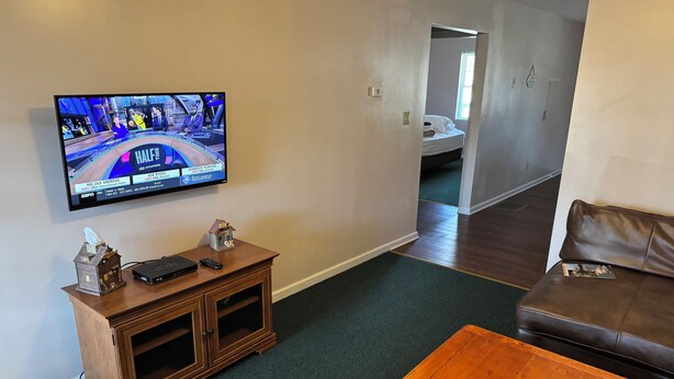 Un salon avec un téléviseur diffusant un match de sport, un meuble en bois, un canapé en cuir marron et une table basse en bois.