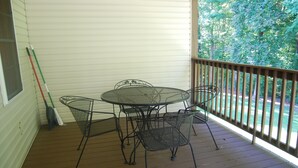 Une terrasse extérieure couverte, avec une table et des chaises en métal, un balai appuyé contre le mur, et une vue sur un paysage verdoyant.