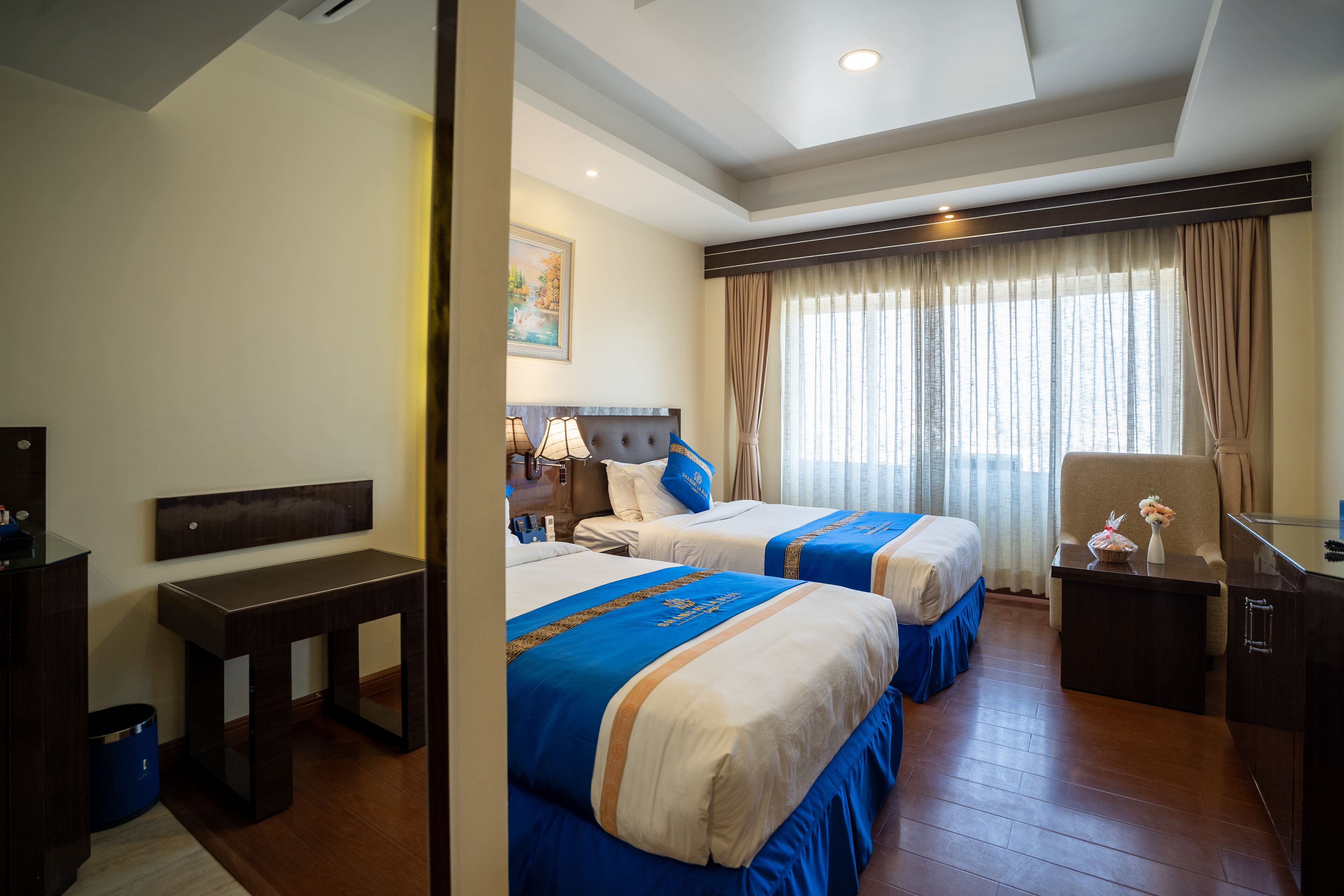 A hotel room with two beds, a desk, a chair, and a large window with curtains.