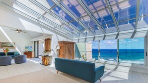 Un vaste salon doté d’un grand plafond en verre, d’un canapé bleu et offrant une vue sur l’océan.