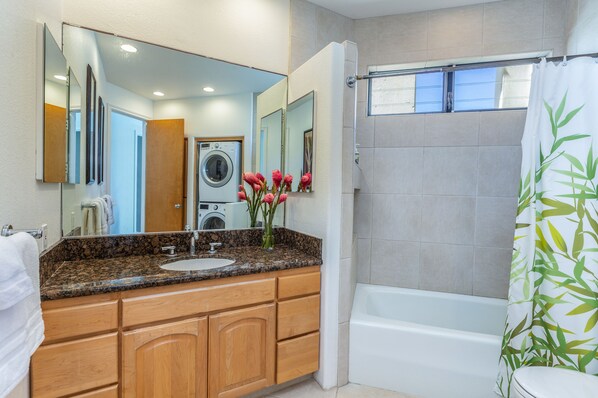 Une salle de bain avec un lavabo sur un plan de travail en granit, des meubles en bois, un grand miroir, une douche avec un rideau de douche en bambou et une fenêtre au-dessus de la douche.
