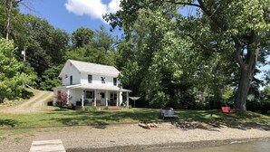 A white house with a porch, a dock extending into a body of water, and a large tree to the right. - Year round lakefront house on Keuka Lake (Hammondsport)
