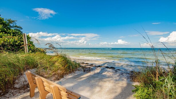 A bench on a sandy path beside a beach with clear blue water and a partly cloudy sky. - Charming Island Escape with Private Pool and Screened Lanai! (Anna Maria)