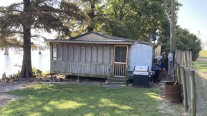 A small wooden cabin with a porch, a grill, and a fenced area with a pond in the background.