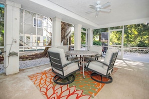 An outdoor patio with a round table and four chairs, a ceiling fan, and a view of a house and trees.
