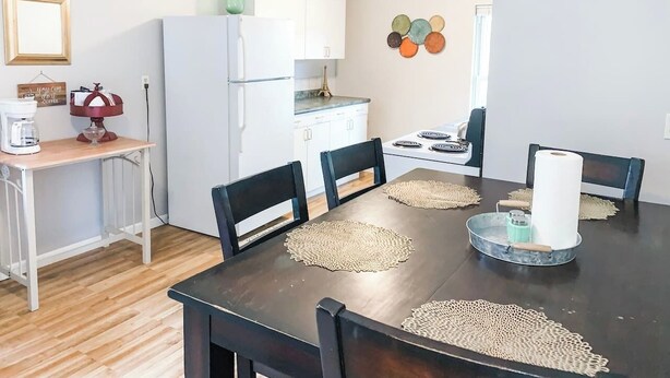 Un comedor con una mesa de madera oscura, cuatro sillas y un objeto decorativo en el centro. Se ve un refrigerador blanco y una cocina con estufa y armarios al fondo.