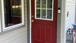 Una puerta roja con un ventanal de marco blanco y un pequeño espacio exterior con una silla de metal y una mesa.