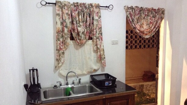 A small kitchen with a sink, cabinets, and a countertop. There is a window with a curtain, and a door leading to an outdoor area. - Porsche Place in San Fernando (Marabella)