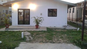 A small, single-story house with a white exterior, a black door, and two windows. There are two potted plants in front. - Porsche Place in San Fernando (Marabella)
