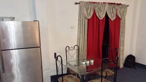 A compact room with a refrigerator, a glass table with chairs, and a window with red curtains. - Porsche Place in San Fernando (Marabella)