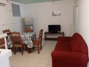 A living room with a red sofa, a dining table with chairs, a TV on a stand, and a refrigerator.