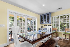 A dining area with a glass-top table, white chairs, and a view through large windows.