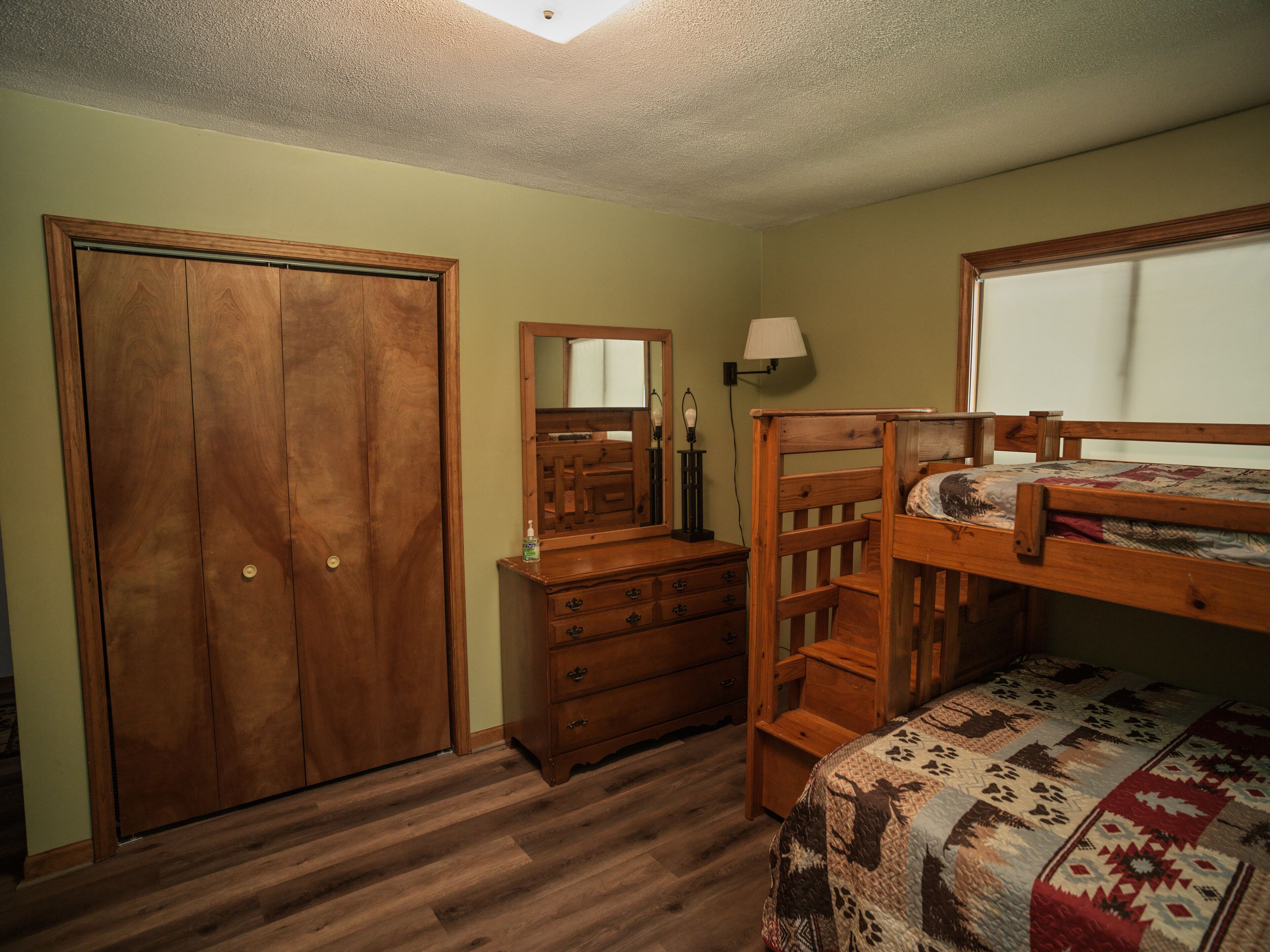 A bedroom with a wooden wardrobe, a dresser, a bed with a quilt, and a window.