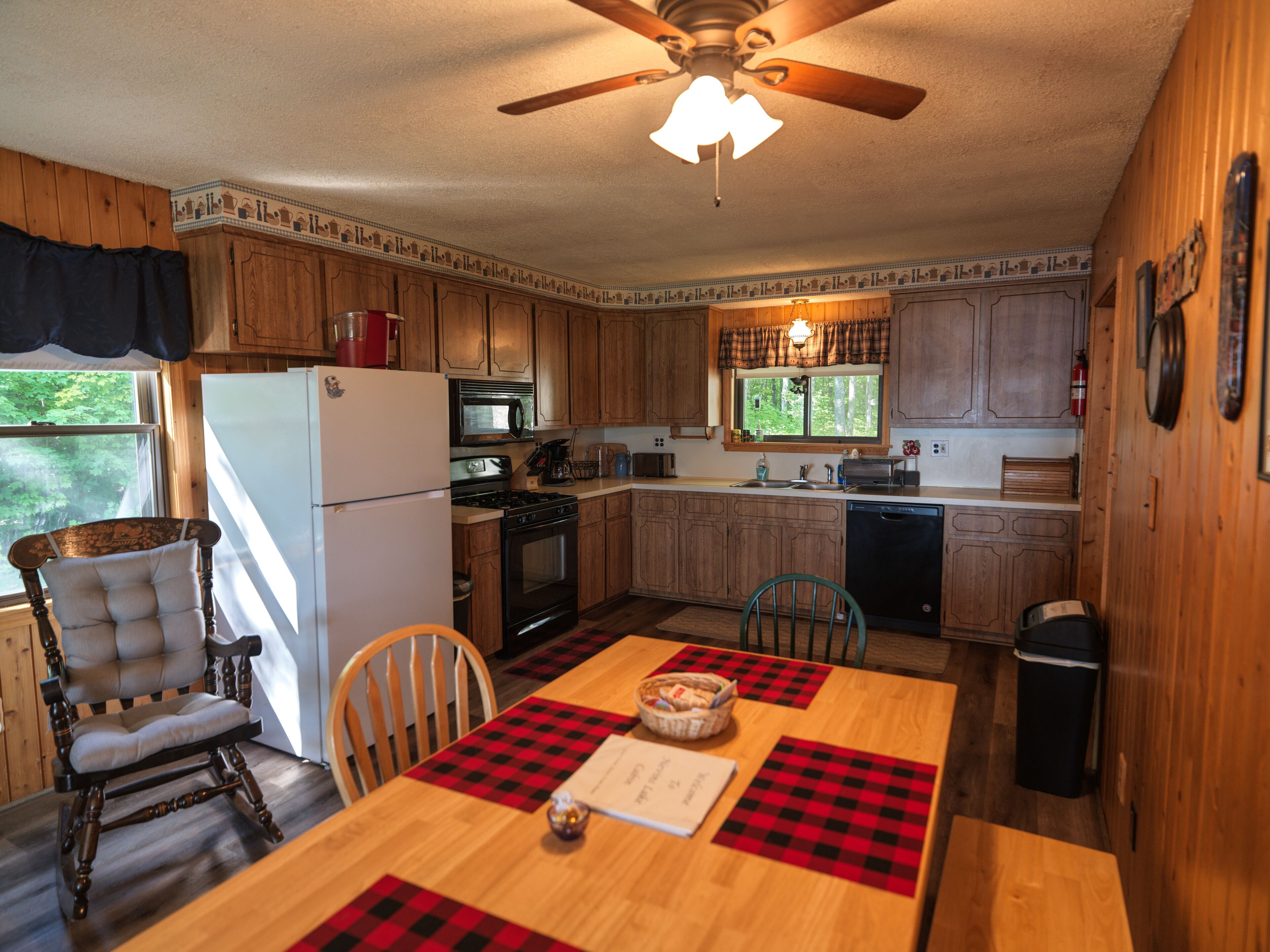 A compact kitchen with wooden cabinets, a refrigerator, stove, microwave, and a dining table with checkered placemats.