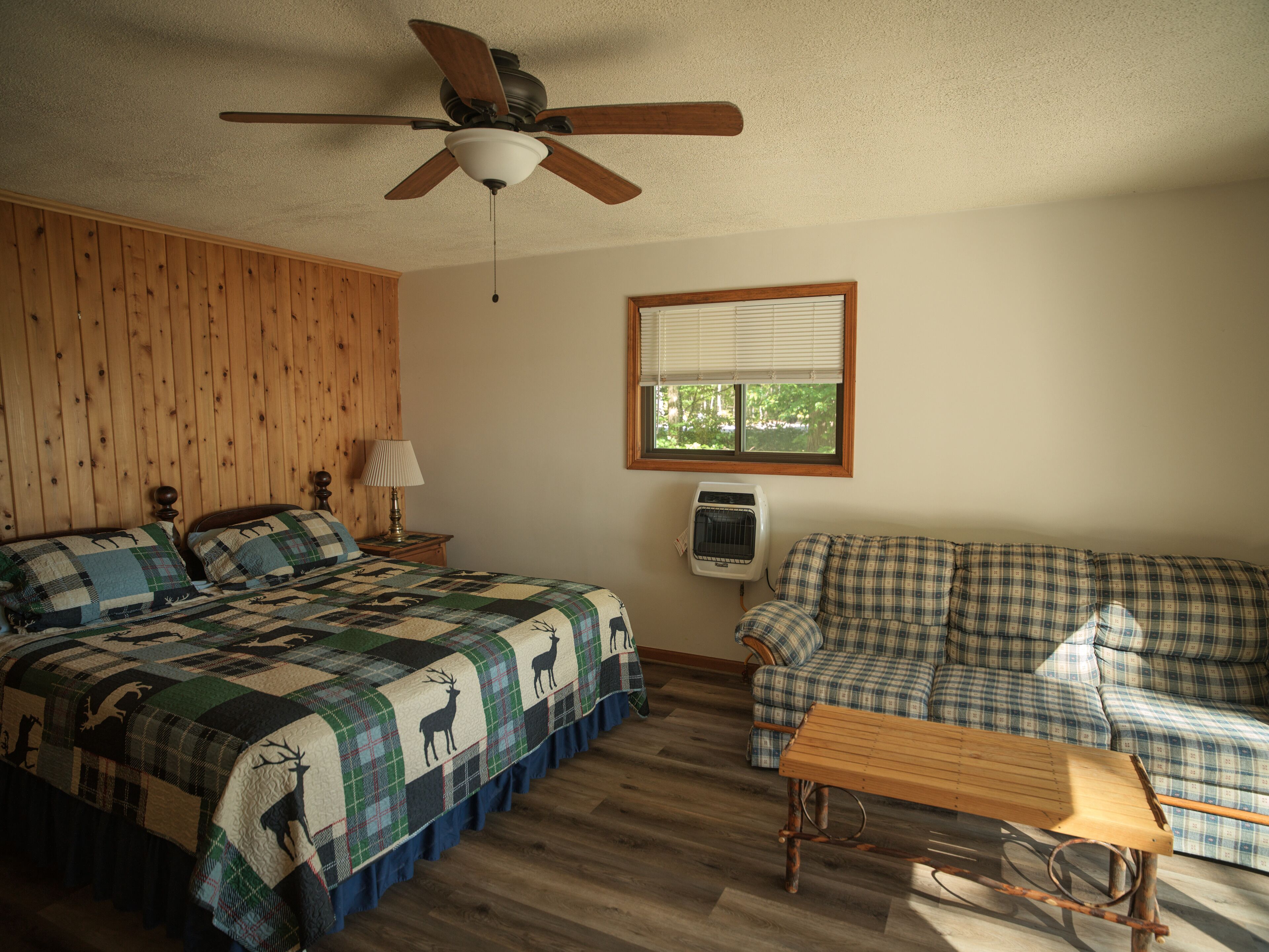 A room with a bed, a wooden ceiling fan, a window with blinds, a wall-mounted heater, and a plaid-patterned sofa.