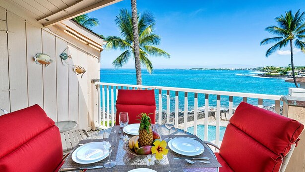 Mesa para dos con cojines rojos, una piña y una flor, en un balcón con vista a la playa.