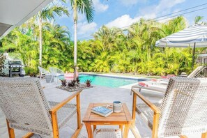 A poolside area with wicker chairs, a wooden table, and a view of a swimming pool surrounded by palm trees. - 529 Whispering Palms Oasis | Private Heated Pool (Naples)