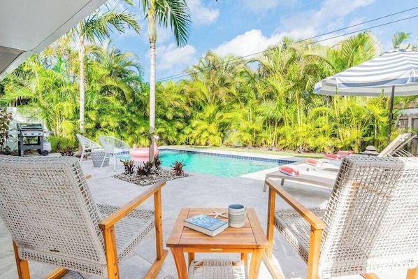 A poolside area with wicker chairs, a wooden table, and a view of a swimming pool surrounded by palm trees.