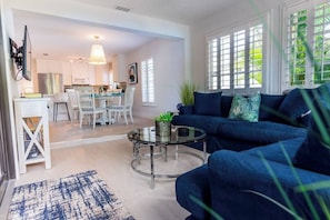 A modern living room with a blue sofa, a glass coffee table, and a dining area with white chairs.