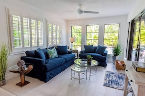 A spacious living room with a sectional sofa, two armchairs, a glass coffee table, and a ceiling fan. Large windows provide ample natural light and views of greenery outside.