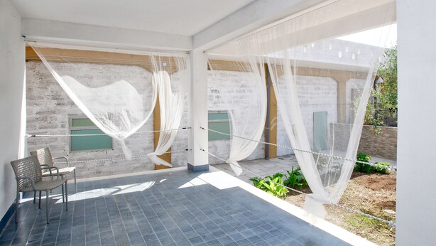 A modern room with a tiled floor, a bed with white curtains, and a view of a brick wall and greenery outside. - Villa al Mare B&B (Giovinazzo)