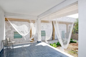 A modern room with a tiled floor, a bed with white curtains, and a view of a brick wall and greenery outside. - Villa al Mare B&B (Giovinazzo)