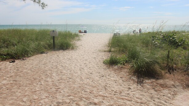 A sandy beach path leading to the ocean with a signpost and beach chairs.