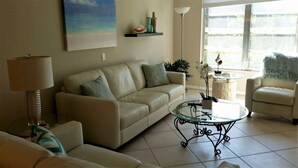A living room with a beige sofa, a glass coffee table, and a lamp. There is a painting on the wall and a window with a view of greenery.