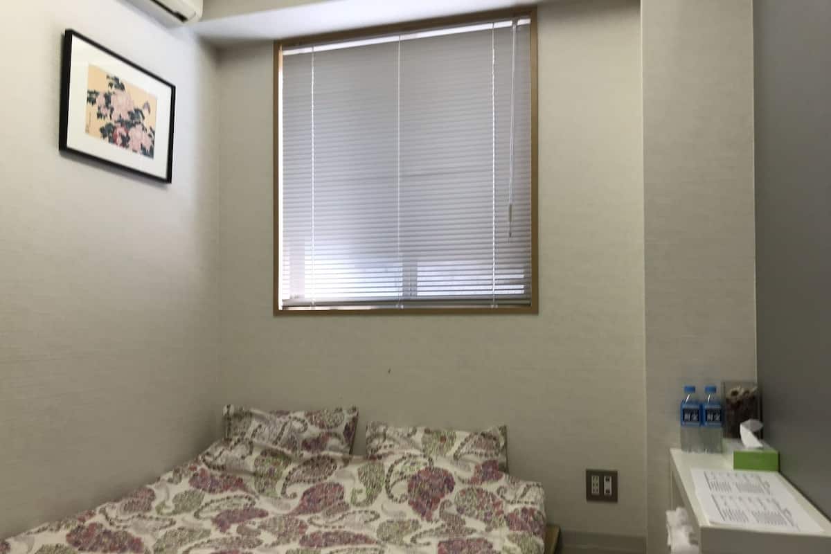 A bedroom with a bed, a window with blinds, a small table with a tissue box, and a picture on the wall.