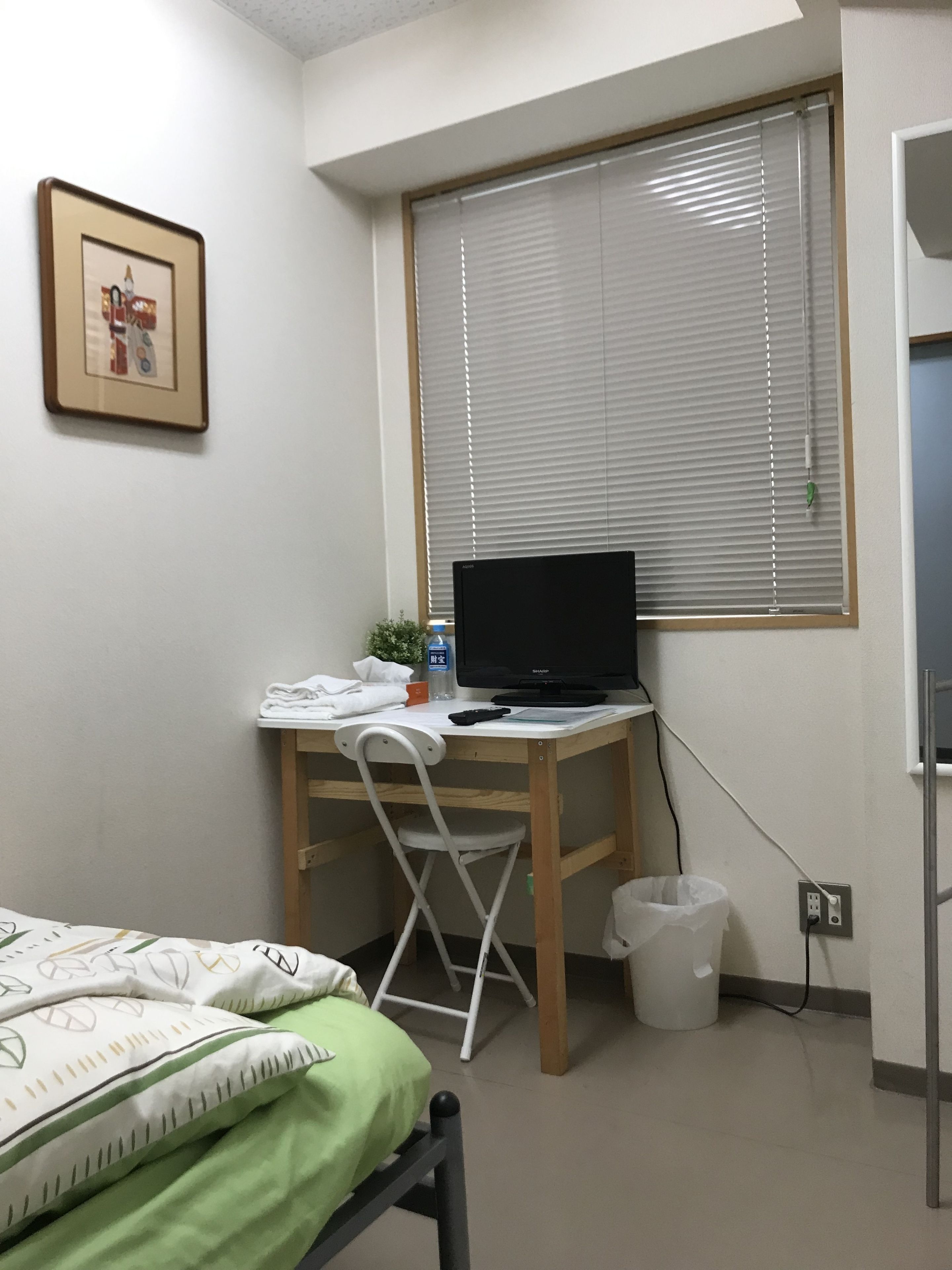 A bedroom with a bed, a desk with a computer, a chair, a window with blinds, a picture on the wall, and a trash bin.
