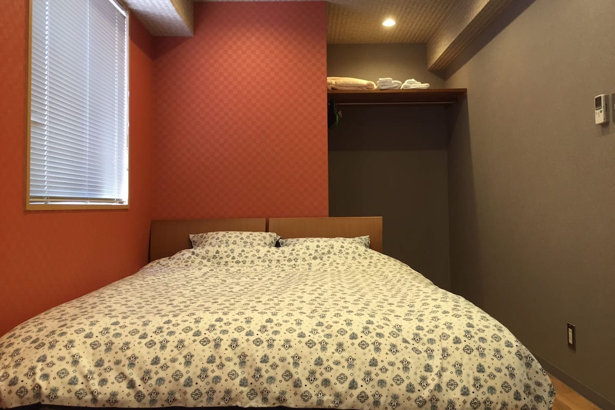 A bedroom with a bed, a window with blinds, a closet with towels, and a ceiling with recessed lighting.