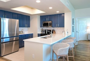 A modern kitchen with blue cabinets, a stainless steel refrigerator, a microwave, and a white island with bar stools. - Hardwood floors & private balcony overlooking lagoon pool! (Destin)