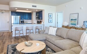 A modern living room with a beige sofa, a round coffee table, and a kitchen area with blue cabinets and a bar stool. - Hardwood floors & private balcony overlooking lagoon pool! (Destin)